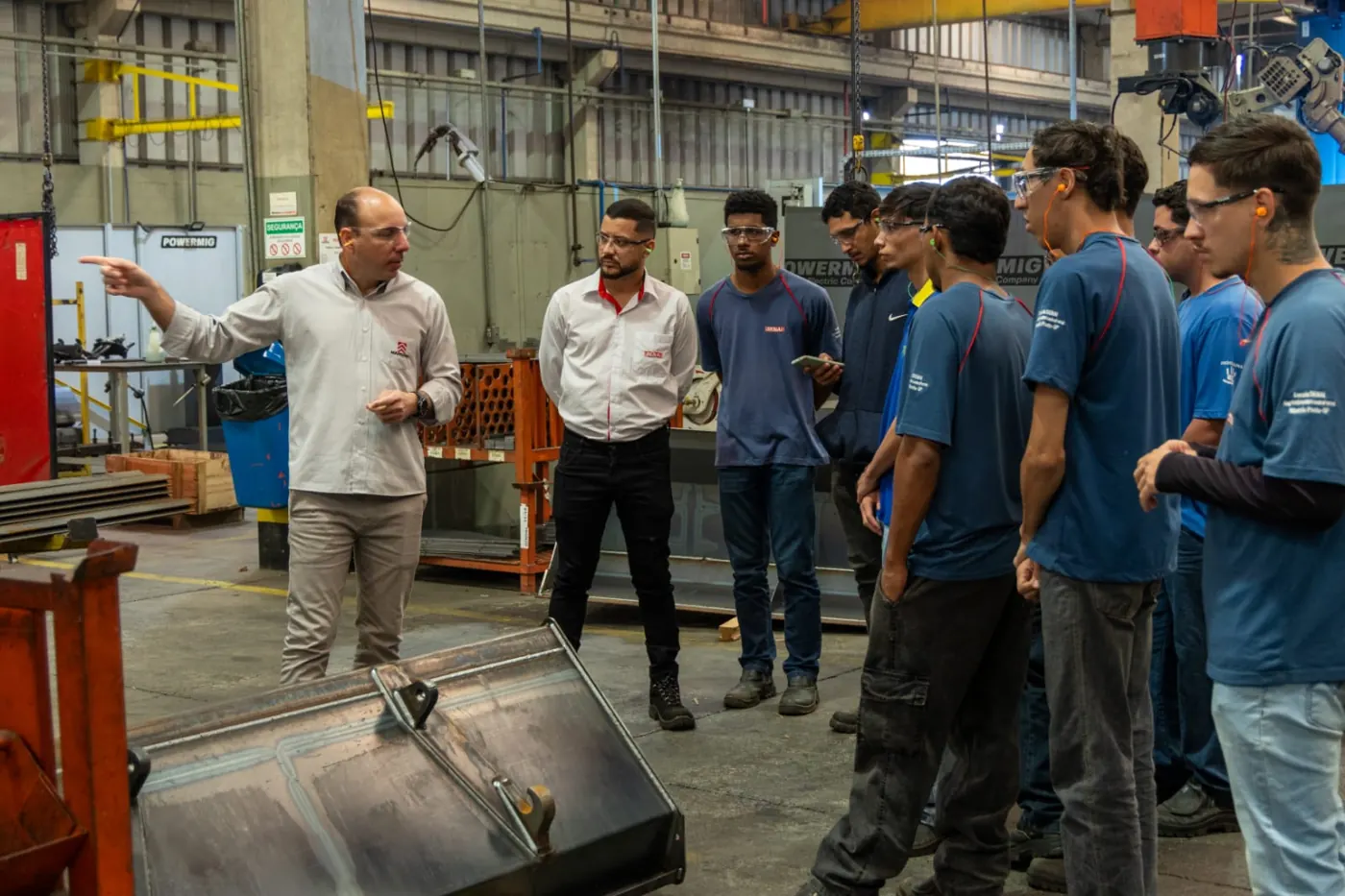 Instrutores e alunos do SENAI, com óculos e protetores auriculares, em visita técnica a uma área de soldagem industrial